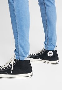 Zapatillas altas de lona negra con suelas de goma blanca y cordones. Combinadas con unos vaqueros ajustados de color azul claro con dobladillos deshilachados.