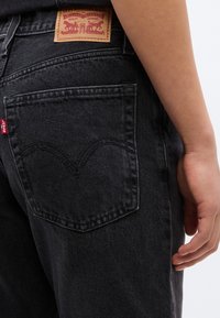 Κοντινό πλάνο στην πίσω τσέπη μαύρου τζιν Levi’s με δερμάτινη ετικέτα μάρκας και κόκκινη ταμπέλα, δίπλα σε χαλαρό χέρι ενός ατόμου.