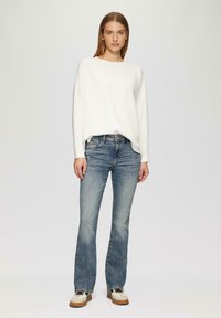 Witte lange mouwen top, relaxte pasvorm, gecombineerd met lichtblauwe flared jeans. Model draagt witte en zwarte sneakers met een bruine zool.