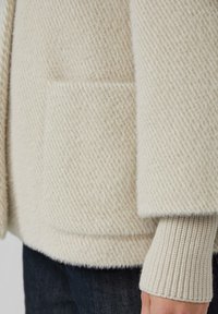 Crèmekleurige gebreide cardigan met een houndstoothpatroon, voorzien van een voorzak en ribgebreide manchetten. Zacht gebreid stofdetail zichtbaar.