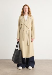 Beige langer Mantel mit breiten Revers und Gürtel, kombiniert mit dunklen Schlagjeans und weißen Sneakers. Trägt eine schwarze Tote-Bag. Glatte Stoffstruktur.