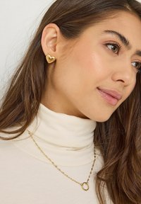 Femme portant des boucles d'oreilles en forme de cœur en or et un collier en chaîne doré avec un pendentif circulaire, vêtue d'un haut à col roulé crème.
