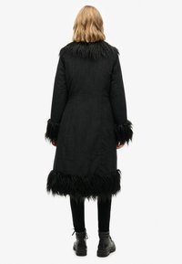 Zwarte jas met nepbontafwerking bij de kraag en manchetten. Lang model, met een gestructureerde stof en een rechte snit. Gecombineerd met zwarte broek en schoenen.