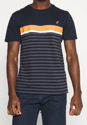 Tmavá osoba s tmavě modrým tričkem s oranžovými a bílými vodorovnými pruhy a malým žlutým ptáčkem na hrudi.