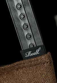 Brown textured fabric with a black plastic strap, featuring circular accents. A black label with "Reell" in white cursive is visible.