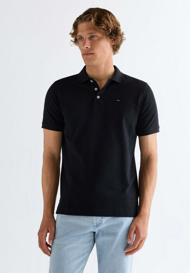 Mand med krøllet hår iført en sort kortærmet polo-shirt og lyseblå jeans, stående mod en ensfarvet hvid baggrund.