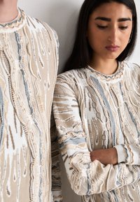 Două persoane poartă pulovere tricotate texturate în nuanțe de bej și crem, cu accente de albastru. Designul prezintă modele complicate și mansete ribbate.