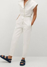 Mono blanco con mangas cortas, botones a presión, cinturilla elástica y bolsillos laterales, combinado con sandalias de tiras azul marino.