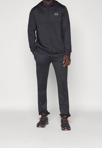 Homme portant un sweat à capuche noir avec fermeture éclair et un pantalon de jogging noir assorti, ainsi que des chaussures de course noires et orange, devant un fond uni.