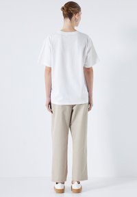 Femme avec les cheveux en chignon portant un tee-shirt blanc, un pantalon large beige, des baskets blanches et des boucles d'oreilles en argent, se tenant devant un mur blanc.