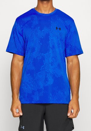 Mężczyzna ubrany w niebieską koszulkę Under Armour z krótkim rękawem w wzory oraz czarne spodenki, stoi na jednolitym tle.