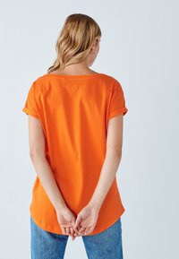 Oranžové krátké tričko s kulatým výstřihem a volným střihem. Materiál se zdá být měkký, s vyhrnutými rukávy a zakřiveným lemem.