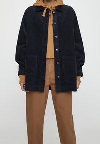 Chaqueta de pana de gran tamaño en color azul marino con cierre de botones, cuello con muesca y dos bolsillos frontales, superpuesta a una sudadera marrón y pantalones beiges.