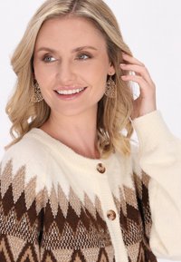 Cardigan crema con motivo geometrico marrone, maglia texturizzata e bottoni grandi. Orecchini a pendente dorati con design intricati.