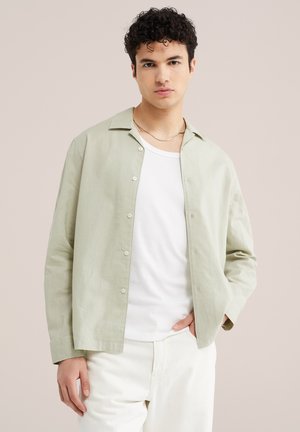 Jeune homme aux cheveux bouclés portant une chemise verte claire boutonnée par-dessus un t-shirt blanc et un pantalon blanc, posant sur un fond beige.