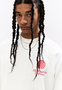 White long-sleeve shirt with red "Alternative Culture" logo. Model wears layered silver and pearl necklaces, and has long twisted hair.