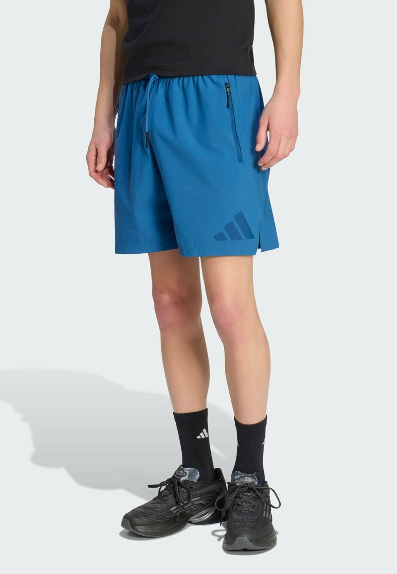 Pantalones cortos deportivos azules con cintura elástica, bolsillos laterales con cremallera y un logo azul oscuro en el lado inferior izquierdo. Calcetines y zapatos negros.