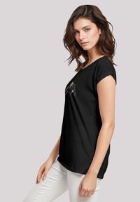 Zwarte t-shirt met een grafisch ontwerp op de voorkant, ronde halslijn en korte opgerolde mouwen. Gemaakt in combinatie met witte broeken.