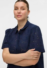 Blusa blu navy con motivi floreali ad occhio, maniche corte a sbuffo e chiusura con bottoni. Tessuto morbido con finitura texturizzata.