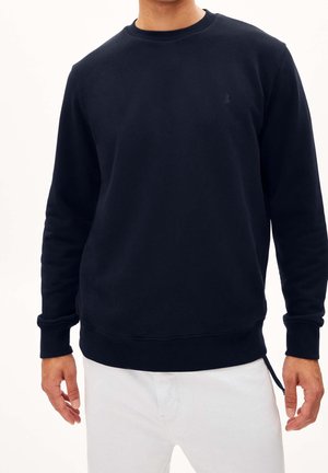 Mężczyzna ubrany w gładką, czarną, długorękawową bluzę z prążkowanymi mankietami, w połączeniu z białymi spodniami na jednolitym tle.