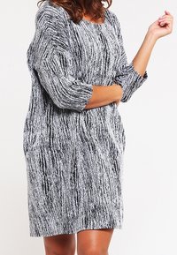 Femme portant une robe longue à manches longues arrivant aux genoux, avec des rayures verticales texturées noires et blanches, debout les bras croisés.