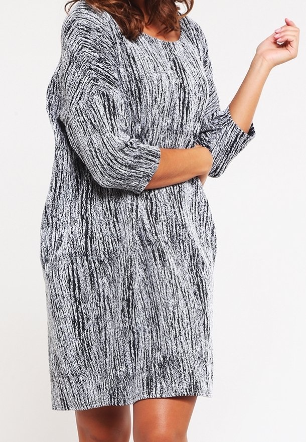 Femme portant une robe longue à manches longues arrivant aux genoux, avec des rayures verticales texturées noires et blanches, debout les bras croisés.