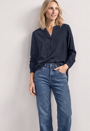 Une femme porte un chemisier bleu marine à manches longues et un jean bleu. Elle se tient debout, une main dans une poche, devant un mur clair et uni.