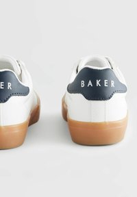 Baskets blanches avec une finition texturée, un onglet de talon bleu marine et le texte "BAKER". Elles présentent une semelle en caoutchouc et un design arrondi avec des coutures contrastées.