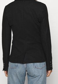 Osoba v přiléhavém černém saka s zapnutými manžetami a světle modrými džíny, viděná zezadu na jednobarevném pozadí.