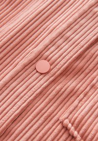 Peach ribbed fabric with vertical pleats, featuring a round button closure and a small pocket detail on the side. Soft texture.