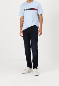 Camiseta de algodón de color azul claro con una franja horizontal roja y blanca y el logo "TOMMY HILFIGER"; combinada con jeans de mezclilla azul oscuro y zapatillas blancas.