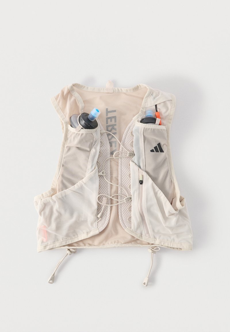 Chaleco de hidratación color beige claro con paneles de malla, bolsillos frontales que contienen dos botellas de agua, cordones ajustables y un pequeño bolsillo con cremallera.