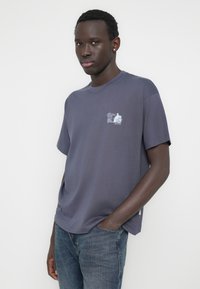 Σκούρο γκρι βαμβακερό t-shirt με κοντά μανίκια, στρογγυλή λαιμόκοψη και μικρό γραφικό σχέδιο στο αριστερό στήθος. Κλασική εφαρμογή με λεία υφή.