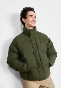 Giacca puffer verde oliva con colletto alto, polsini a coste e un logo nero sul petto, caratterizzata da una trama trapuntata e una vestibilità rilassata.