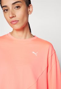 Kortermet, korallfarget treningsskjorte laget av glatt stoff, med en hvit Puma-logo på venstre bryst og rund hals.