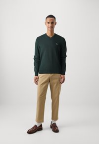 Maglione verde a scollo a V in un materiale morbido, abbinato a chinos beige e mocassini marroni. L'outfit è completato da calzini bianchi.