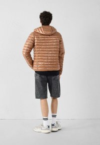 Giacca imbottita marrone con cappuccio, design trapuntato e texture lucida, abbinata a pantaloncini di denim scuro e scarpe sportive con accenti a contrasto.