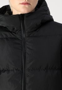 Juodas pūkinis kailis su atsegamu kapišonu, su priekyje esančiu užtrauktuku, išsiuvinėjimu ir siuvinėjimo detalėmis.