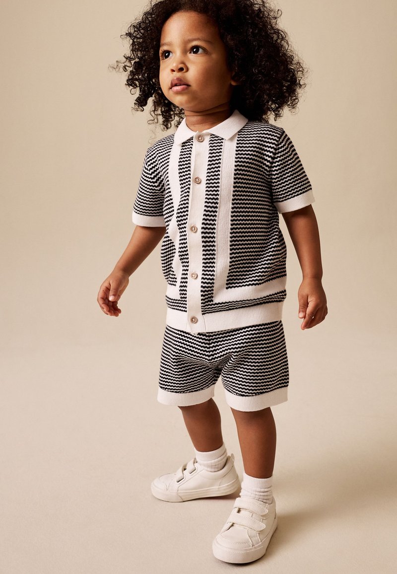Niño pequeño con pelo rizado que lleva una camiseta de manga corta y pantalones cortos con estampado en blanco y negro, calcetines blancos y zapatillas blancas, de pie sobre un suelo beige.