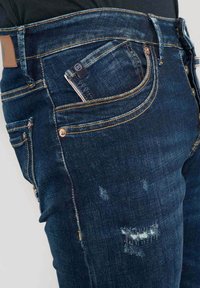 Jeans de mezclilla azul oscuro con parches desgastados, costuras en contraste, cinco bolsillos y un parche de cuero en la parte posterior. Diseño ajustado, cintura de altura media.