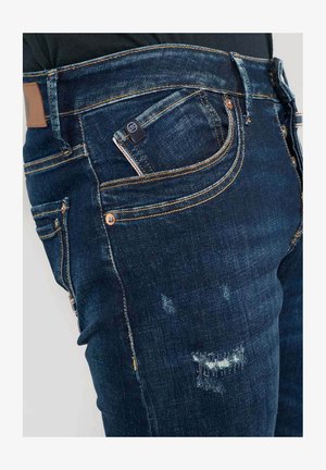 Jeans de mezclilla azul oscuro con parches desgastados, costuras en contraste, cinco bolsillos y un parche de cuero en la parte posterior. Diseño ajustado, cintura de altura media.