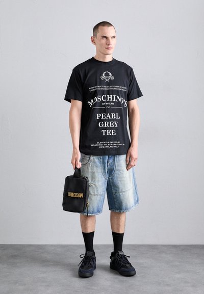 Tricou negru cu text alb, pantaloni scurți din denim cu o nuanță de albastru estompat, șosete negre și adidași negri. Ținând o geantă mică neagră cu text auriu.