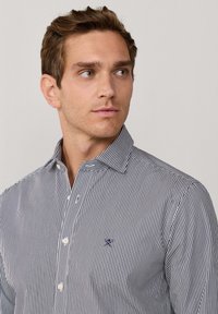 Raitapaita tummansinisessä ja valkoisessa, jossa on napitettava kaulus, pitkät hihat ja pieni brodeerattu logo rinnassa. Sileä kankaan rakenne.