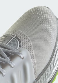 Jasnoszary but sportowy z teksturowanym materiałem wierzchnim, wyściełanym kołnierzem oraz elastyczną podeszwą w zielonych akcentach i odbijającym srebrnym wykończeniem.