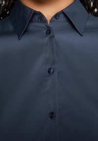 Gros plan d'une chemise bleu marine foncé à col avec quatre boutons visibles sur le devant, portée par une personne à la peau foncée.