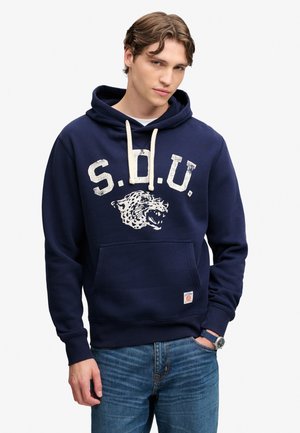 Ung mand iført en navyblå hættetrøje med hvide "S.D.U." bogstaver og en brølende leopardgrafik, kombineret med blå jeans og et armbåndsur.