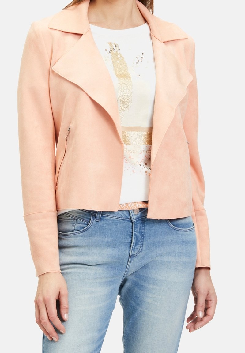 Femme portant une veste en daim rose clair par-dessus un t-shirt blanc à imprimé graphique et un jean bleu clair, vue des épaules aux hanches.