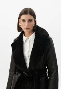 Černý kožený kabát s límcem z umělé kožešiny, dlouhými rukávy a opaskem. Nositelný přes bílou košili s knoflíky. Obsahuje kulaté zlaté náušnice.