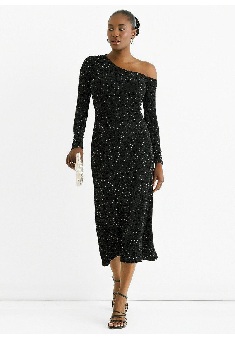Black, form-fitting dress with one shoulder, long sleeve, and white polka dots. Midi length with soft texture, paired with black strappy sandals.