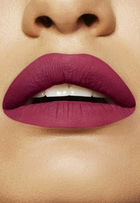 Gros plan de lèvres portant un rouge à lèvres mat rose foncé, légèrement entrouvertes, révélant les dents supérieures avant et la peau texturée autour de la bouche.